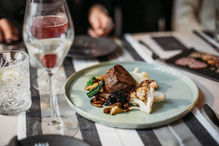Tasty grilled steak served with cauliflower and mushrooms placed on table with wineglass filled with alcohol drink in restaurant on blurred background